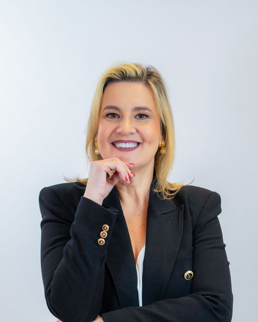 Polly Montarroyos de blazer escuro, sorrindo, com a mão no queixo